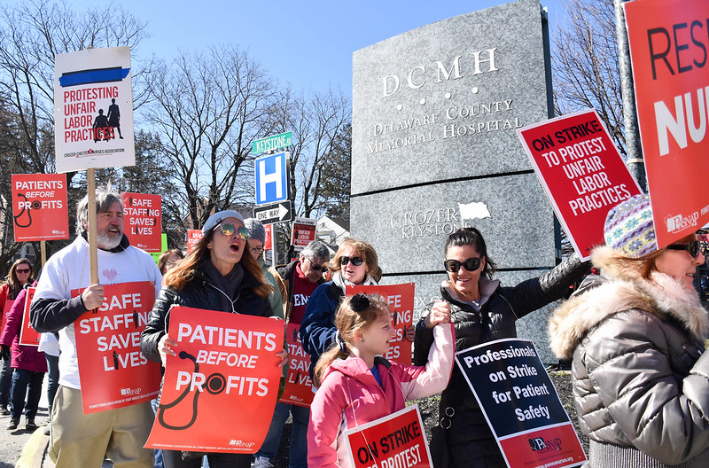 Eleven nurses picket in front of a hospital sign carrying red signs reading Protesting Unfair Labor Practices, Patients Before Profits, Professionals on Strike for Patient Safety.