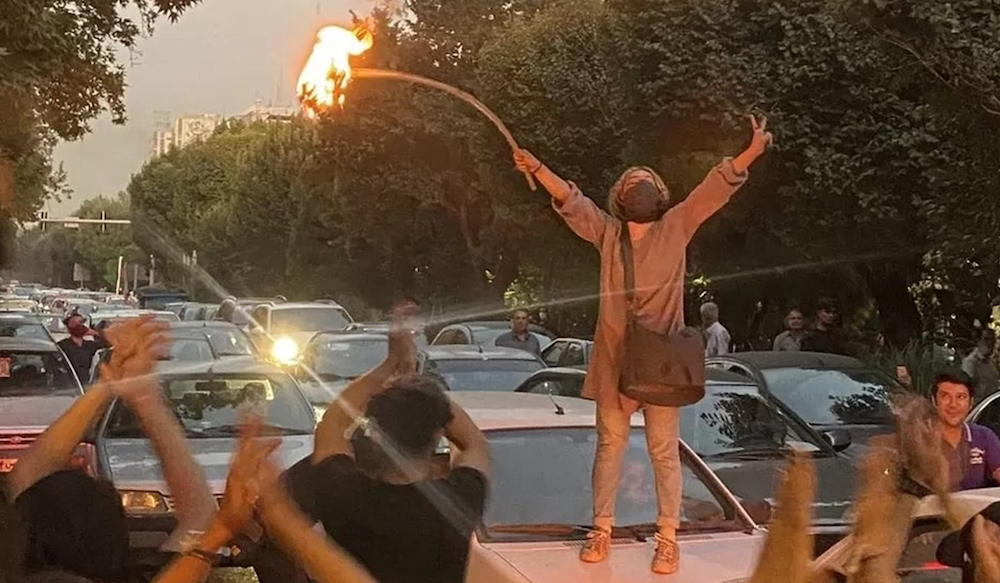 Photo of Iranian protester standing on a car with her hands over head with a torch in one hand and making a victory/ peace sign with the other hand. 