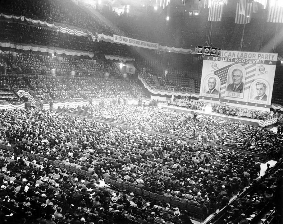 A photograph of a large rally of the American Labor Party, likely at Madison Square Garden in New York City, ahead of the 1940 presidential elections. In addition to the large crowd filling the stadium, behind the stage are three huge images of candidates, under the banner of the American Labor Party. These are: presidential candidate Franklin Delano Roosevelt, vice-presidential candidate Henry Wallace, and New York senate candidate James Mead.