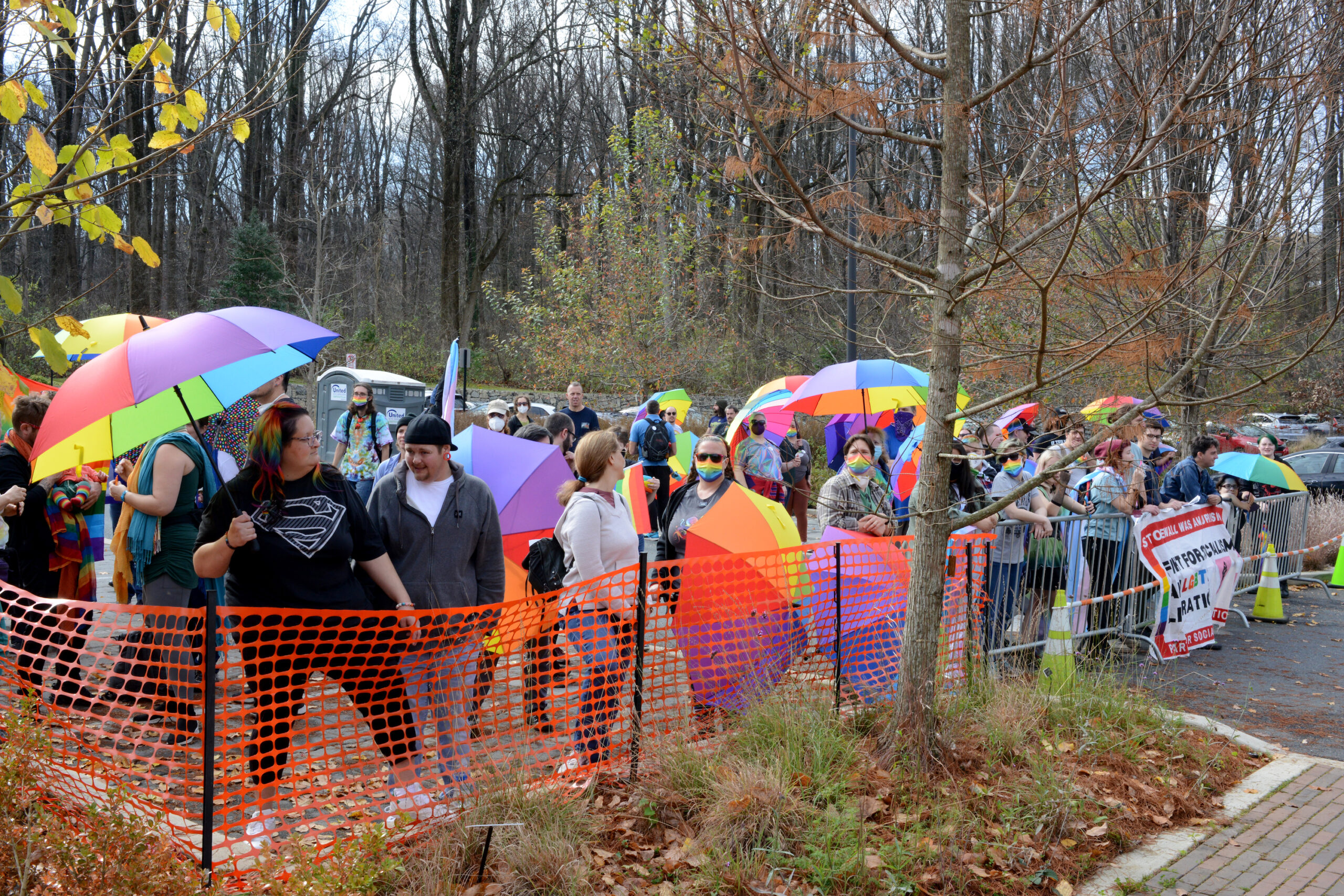 A crowd of about 100 people, many holding rainbow umbrellas, gather behind an orange barricade in a forest. They are in Silver Spring, Maryland defending a Drag Story Hour event from far-right protesters.