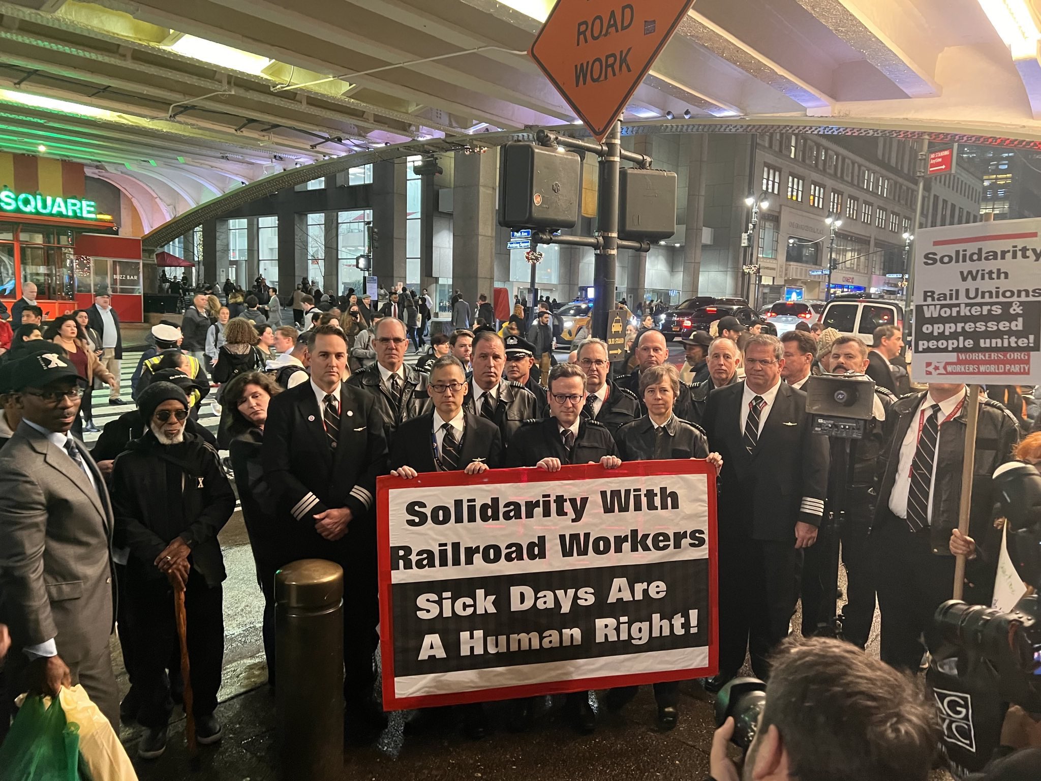 Railroad workers in uniform stand behind a banner that reads, “Solidarity with Railroad Workers Sick Days Are A Human Right!”