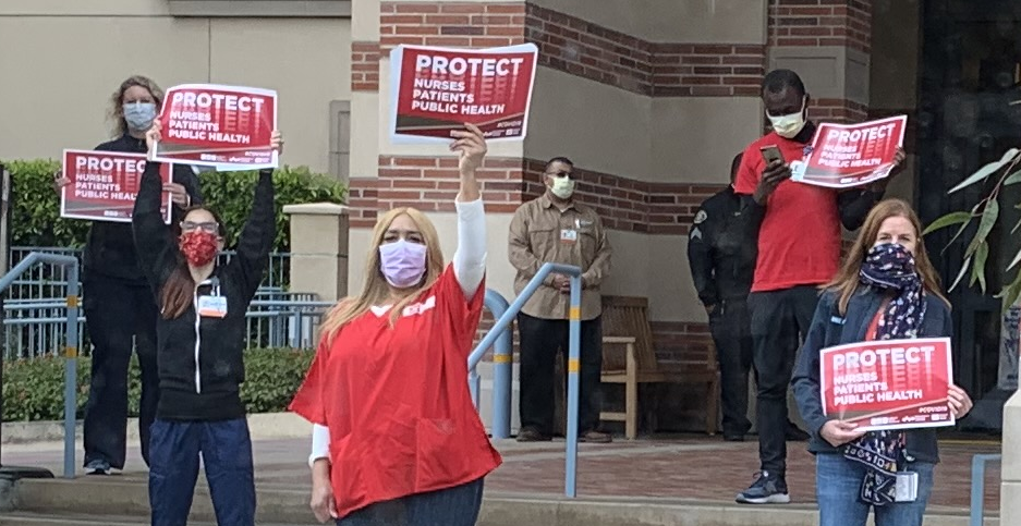Five nurses wearing surgical masks stand distanced from one another on the front steps of a building. They hold up red signs that say: PROTECT. NURSES. PATIENTS. PUBLIC HEALTH. Two security personnel stand in the background.
