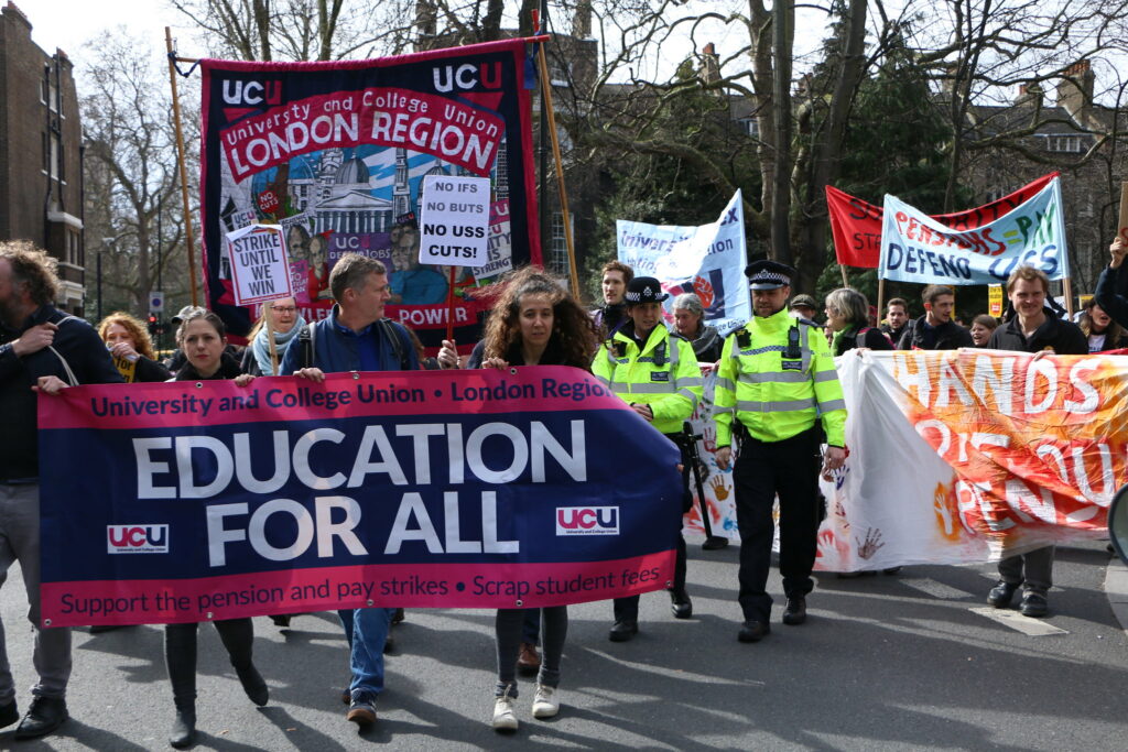 People march down a street holding various signs. The largest sign says ‘University and College Union - London Region, EDUCATION FOR ALL, Support the pension and pay strikes - Scrap student fees’ and is pink and purple, the colors of UCU. Another sign says ‘HANDS OFF OUR PENSION’ in bright red and orange colors, with painted handprints. Two cops walk among the protestors.