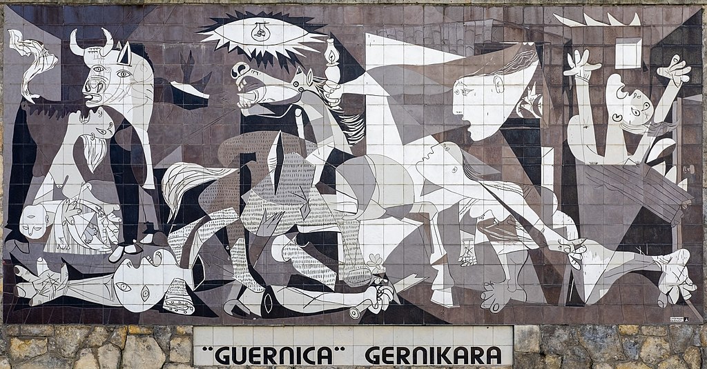 A mural of the painting ‘Guernica’ by Picasso made in tiles and full size. The imagery is black, shades of gray and white. It consists of animals, a screaming woman, a dead baby, a dismembered soldier and flames. Underneath the mural is a sign that says: “GUERNICA” GERNIKARA.