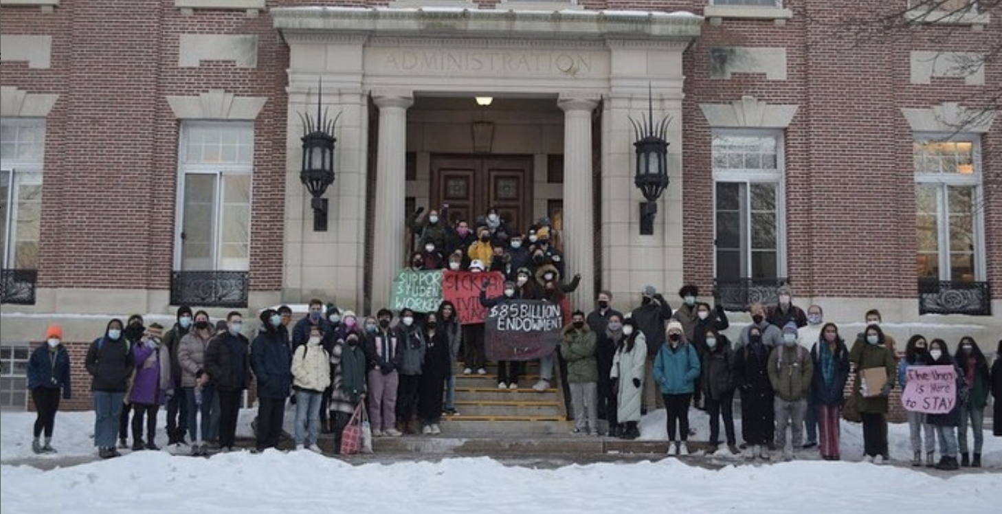 A large group of people, presumably Dartmouth students, pose for a photo in front of a brick building titled ‘ADMINISTRATION.’ Various students pose with a power fist and hold up signs, one reads ‘SUPPORT STUDENT WORKERS,’ another reads ‘SICK DAYS - LIVING WAGES,’ another ‘$85 BILLION ENDOWMENT’ with other writing that is difficult to discern, and another reads ‘The Union is Here to STAY.’ Everyone is dressed for the cold winter, and snow covers the ground.