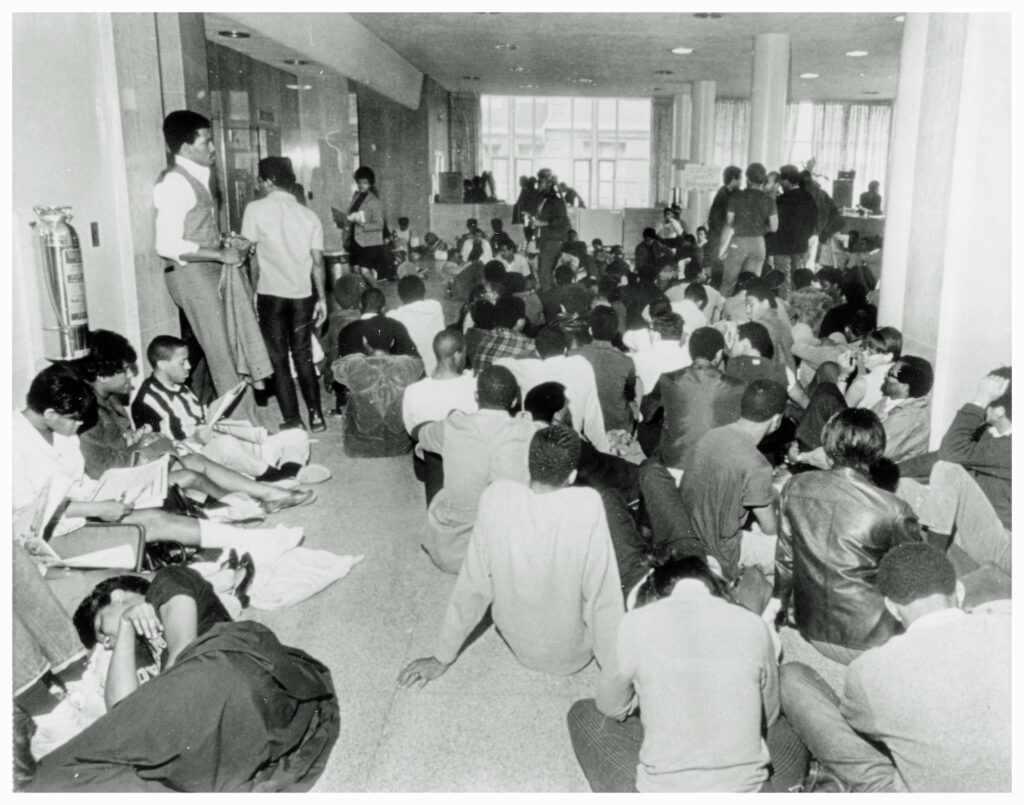 Dozens of students, in a black and white photo, sit, stand, sleep, and talk in the hallway of an administrative building at Howard University.