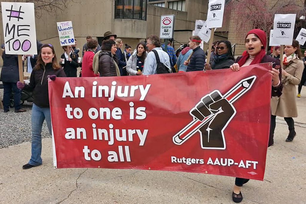 Picketers march carrying a red banner reading An Injury to One is an Injury to All: Rutgers AAUP-AFT. The image is of a fist raised, gripping a pencil.