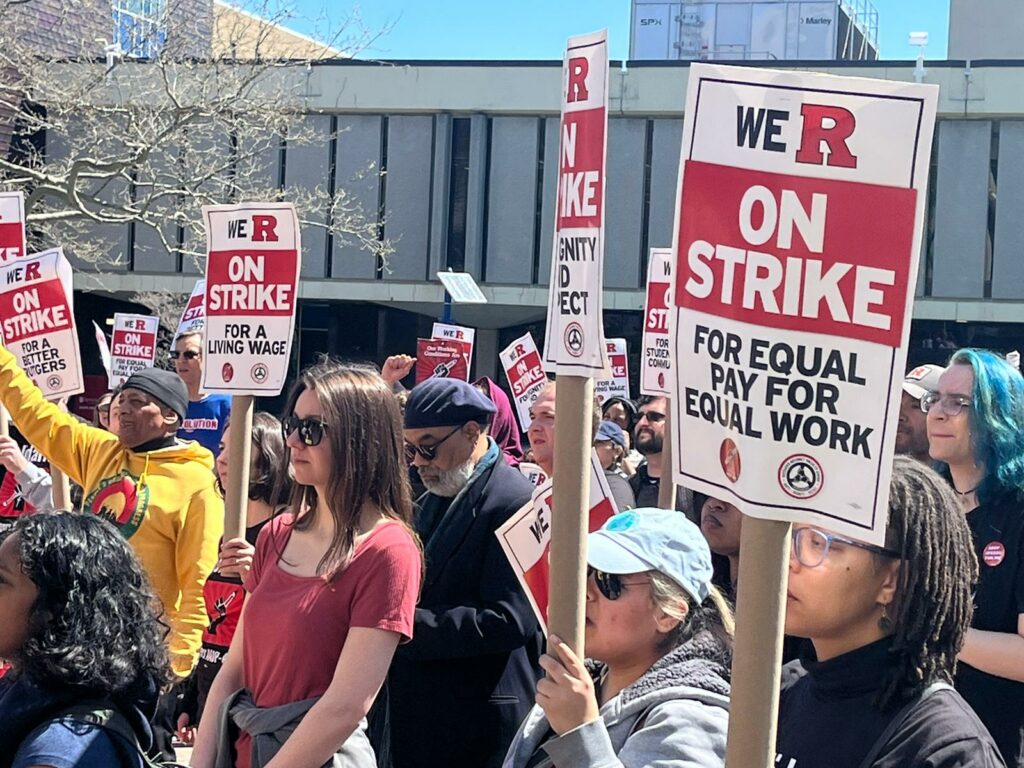 A crowd of demonstrators in front of campus buildings on a sunny day hold signs reading We R on Strike for Equal Pay for Equal Work.