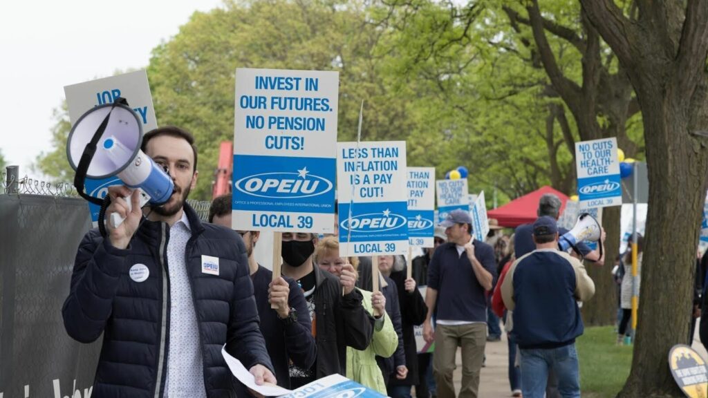 Joe Evica, a bearded white man with short brown hair, is pictured on the left holding up a bullhorn and speaking into it. Joe is followed by a line of workers walking along a tree-lined sidewalk and holding blue and white picket signs that bear the OPEIU Local 39 union logo. The signs feature different slogans, including: “INVEST IN OUR FUTURES, NO PENSION CUTS!” “LESS THAN INFLATION IS A PAY CUT!” and “NO CUTS TO OUR HEALTH-CARE.”