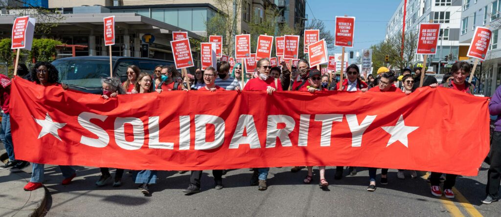 Starbucks workers march behind a giant red banner that says “SOLIDARITY,” holding picket signs with various slogans, including, “DEMAND THAT STARBUCKS REINSTATE ALL WORKERS FIRED FOR ORGANIZING!” and “REBUILD A FIGHTING LABOR MOVEMENT,” and “AN INJURY TO ONE IS AN INJURY TO ALL!”