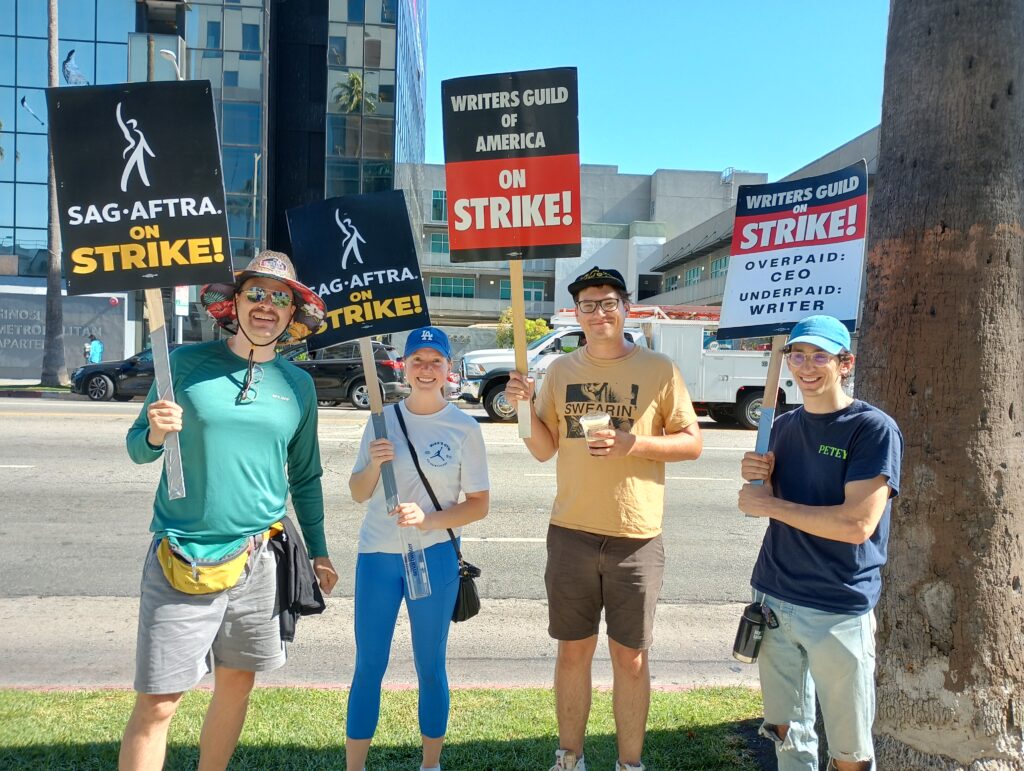 Four people stand with picket signs representing SAG-AFTRA and WGA strikes
