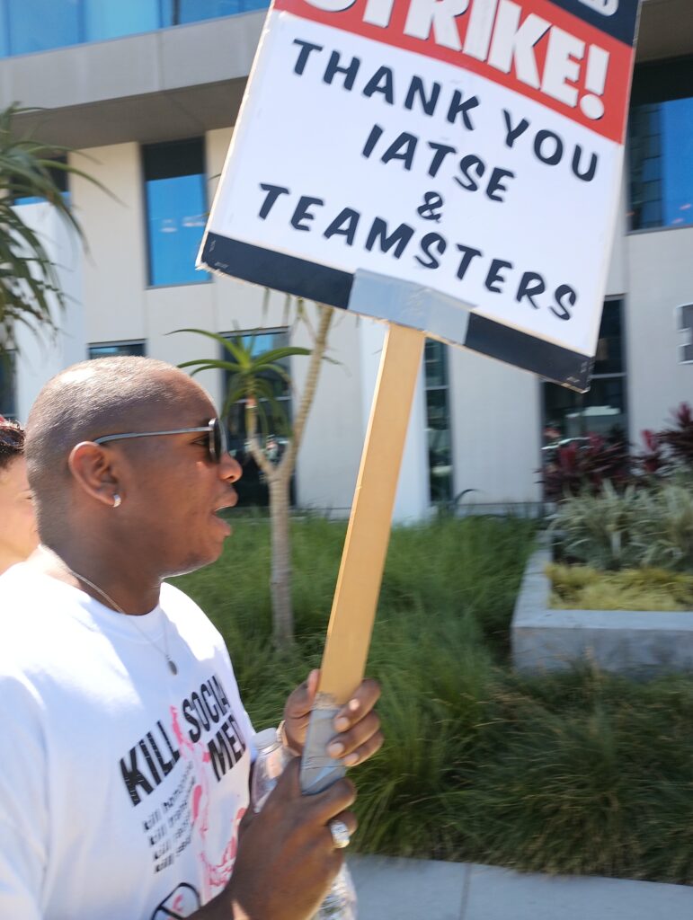 A Black man holds a picket sign in red and blue reading Strike! Thank you IATSE and Teamsters
