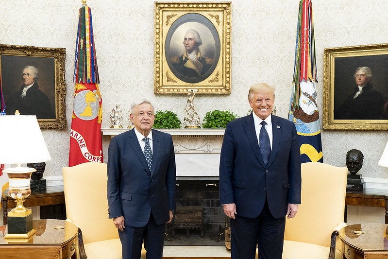 President Andrés Manuel López Obrador is on the left standing next to President Donald Trump on the right in the White House Oval Office. Both men are standing in front of two yellow chairs, a fireplace, military flags representing the Marine Corps and the Navy, and portraits of Presidents James Madison (left) George Washington (center), and Thomas Jefferson (right).