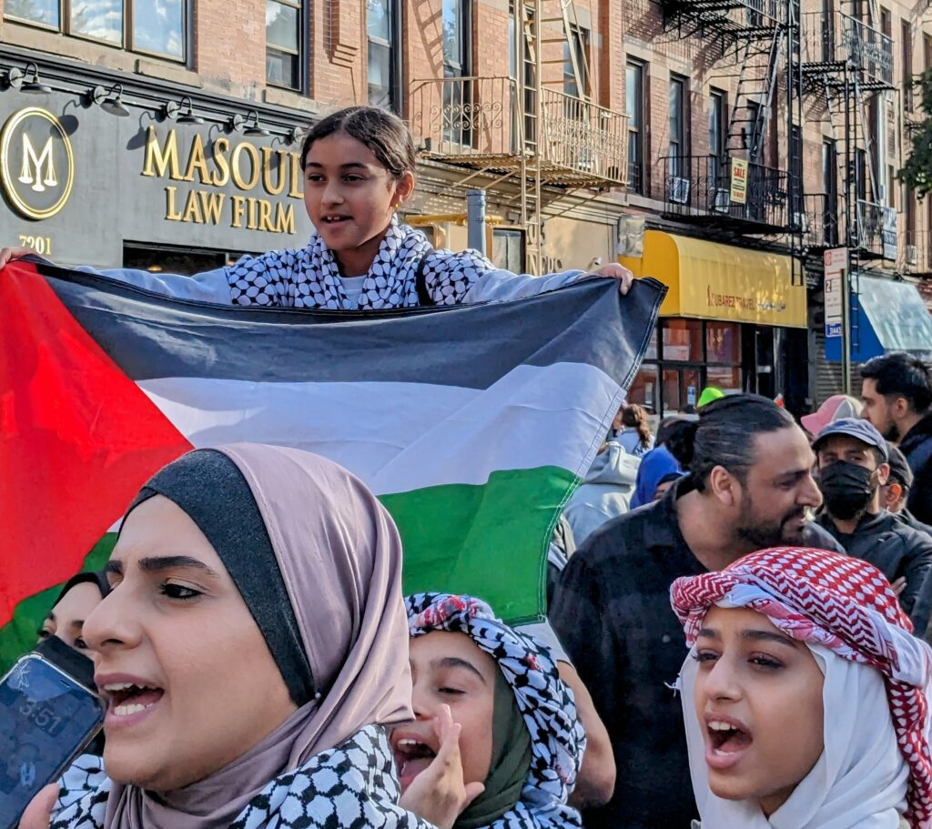 A child rides the shoulders of a marcher in a massive demonstration for Palestine. She is carrying a broad Palestinian flag.