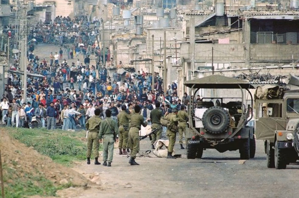 Long shot of a crowd of hundreds of Palestinian massed in a street, facing the camera in multicolored clothes; some in the front rank are throwing rocks. In the foreground are about ten Israeli soldiers dressed in olive drab with backs faced to the camera.
