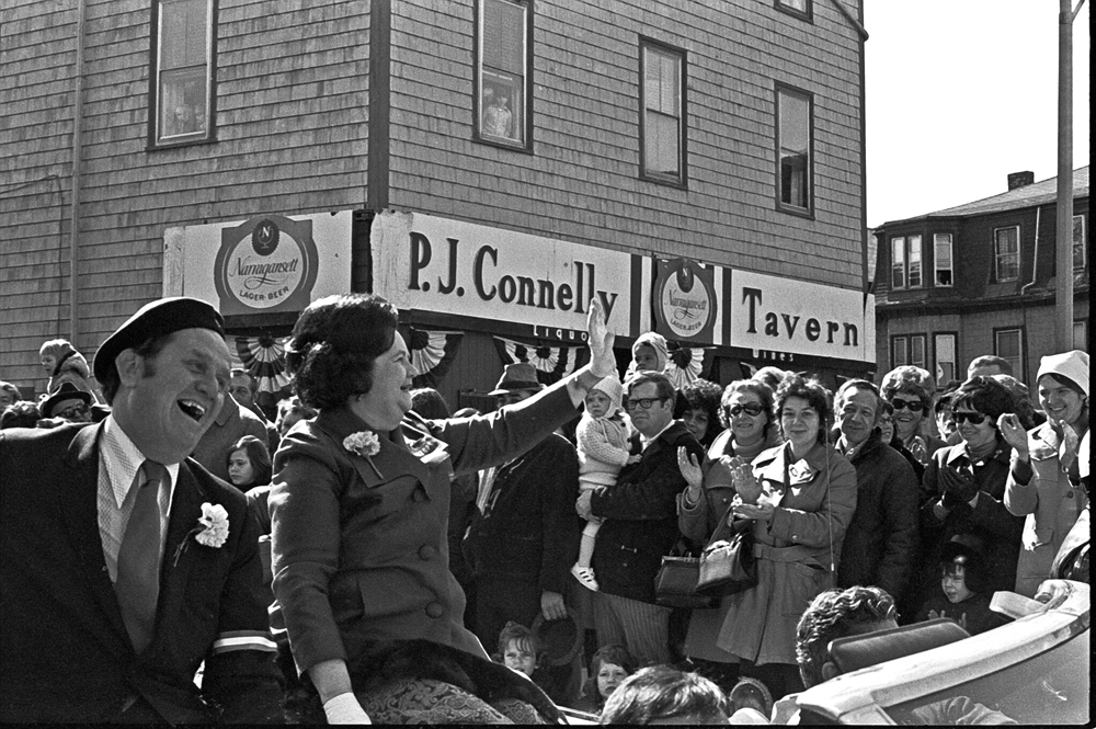  Black and white photo. A middle-aged white women wearing white gloves and a carnation is perched on the back of a convertible car and waves to a crowd that has gathered to watch the parade. Riding next to her is a man with a broad smile and a carnation. The all-white crowd smiles and applauds. Children look on from windows above a building marked with a sign reading, “P. J. Connelly Tavern.”