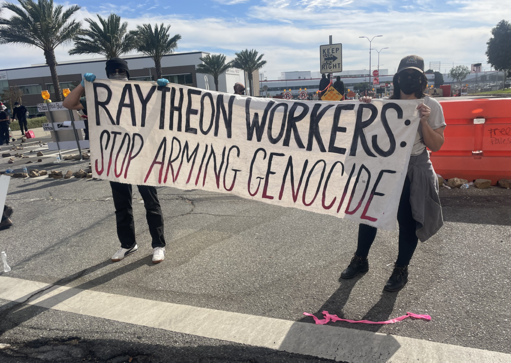 Two masked activists hold a banner, in black and red on white, reading Raytheon Workers: Stop Arming Genocide.