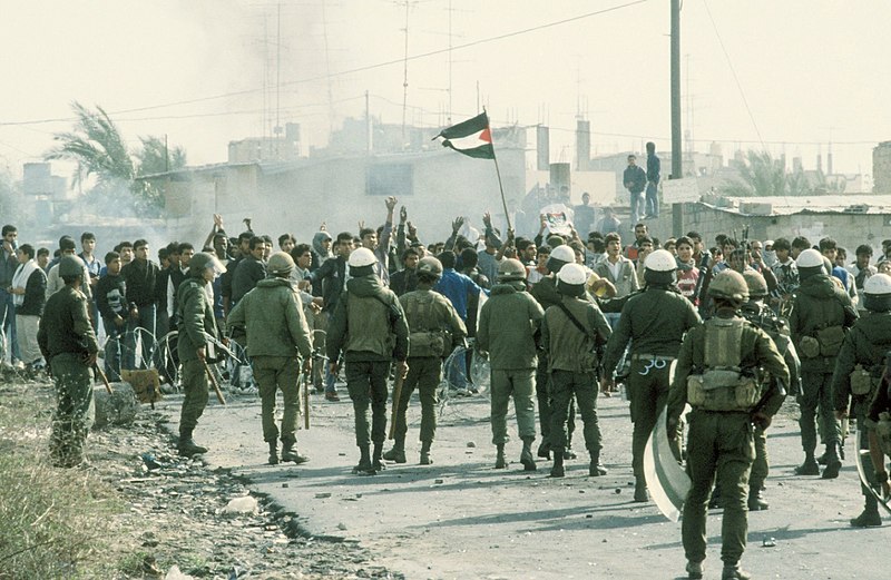 A mass of Palestinian fighters, photographed from behind, enters a smoke-filled urban area.