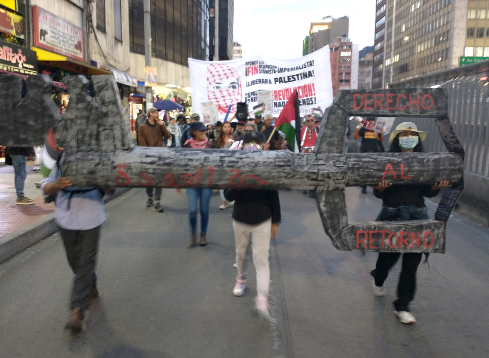 Photo of a street march, apparently in the evening. Two marchers, shot from in front, carry a black and gray papier maché key with the words “Derecho al retorno”—“the right of return.”