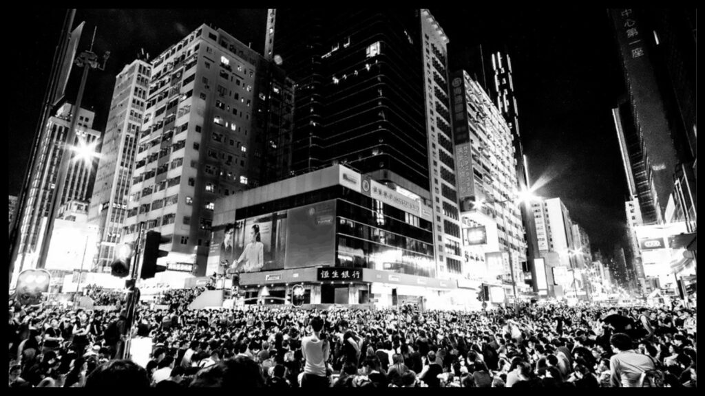 Black and white photo. A view, from slightly above, of a crowd at a large intersection of streets. Modern buildings of ten or more stories tower above, and the packed crowd extends down both of the two intersecting streets as far as the camera can see.