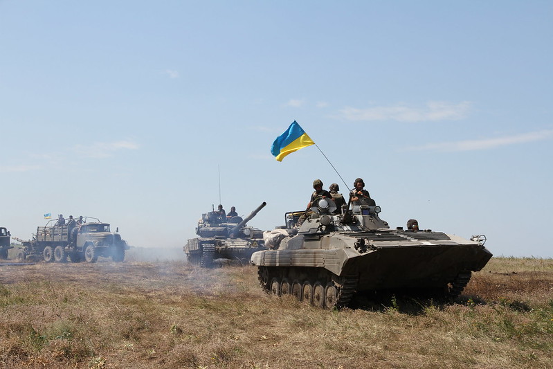 Ukrainian soldiers in two tanks in the front and two trucks in the back with one only slightly in view of the image are shown moving through an open field in eastern Ukraine in July 2014. The Ukrainian flag is proudly displayed on the top of the first tank in the front and on one of the trucks in the back.