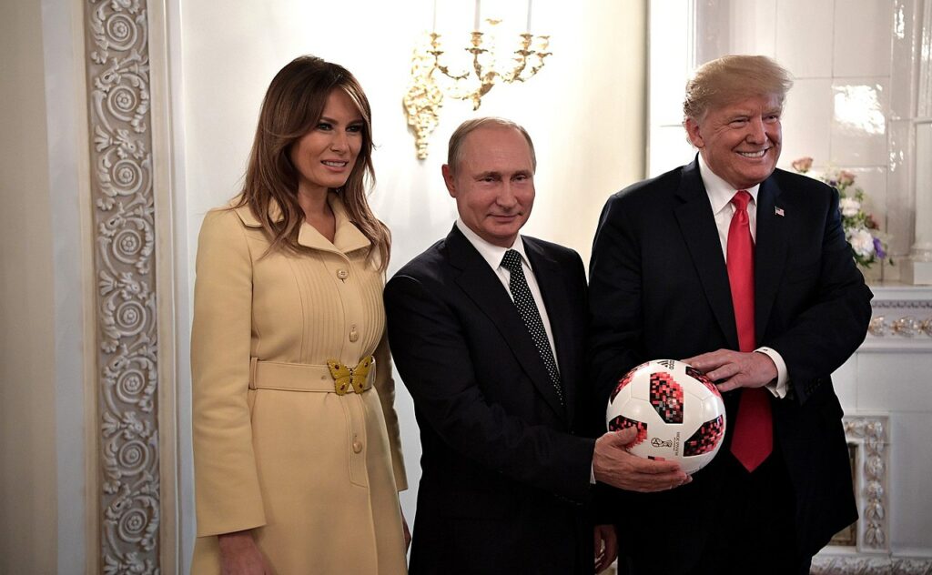 While meeting in Helsinki, Finland, Vladimir Putin in the middle poses in a suit with a slight smirk on his face for a picture along with Melana Trump who is awkwardly smiling on his right wearing a yellow coat with a butterfly belt and Putin is handing an official 2018 FIFA World Cup Adidas soccer ball (football) to Donald Trump on his left who is smiling while wearing a suit with a red tie and a U.S. flag pin.