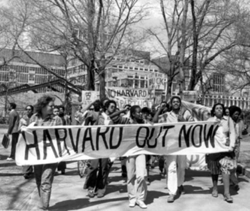A black and white photo of students marching with a banner reading Harvard Out Now