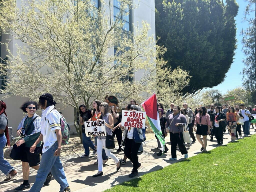 A group of people marches carrying a Palestinian flag and signs reading Zionism is fascism and Israel bombs babies.