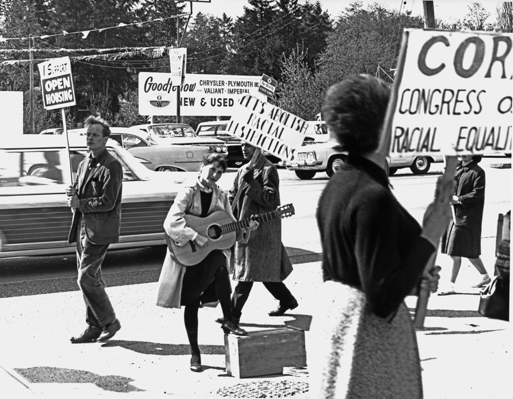 A black and white photo offour activists picketing with signs reading Abolish Rafcial Inspections and CORE: Congress of Racial Equality. One protester is playing a guitar with her foot up on a box.