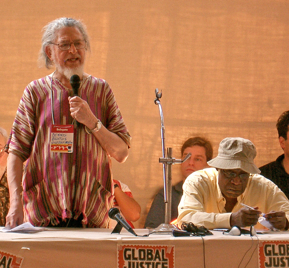An old man in an African cotton tunic speaks into a handheld microphone with other speakers seated nearby. (Photo taken from the audience.)