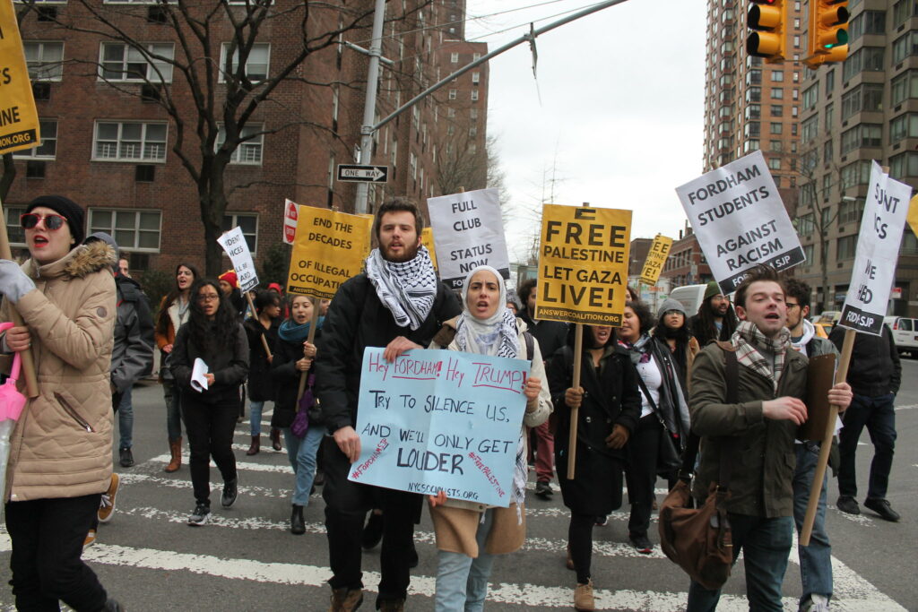 Dozens of protesters march toward the camera across a New York city street. They carry signs reading Try to Silence Us, We will only get louder; free Palestine; let Gaza live. The two activists in the foreground are wearing keffiehs.