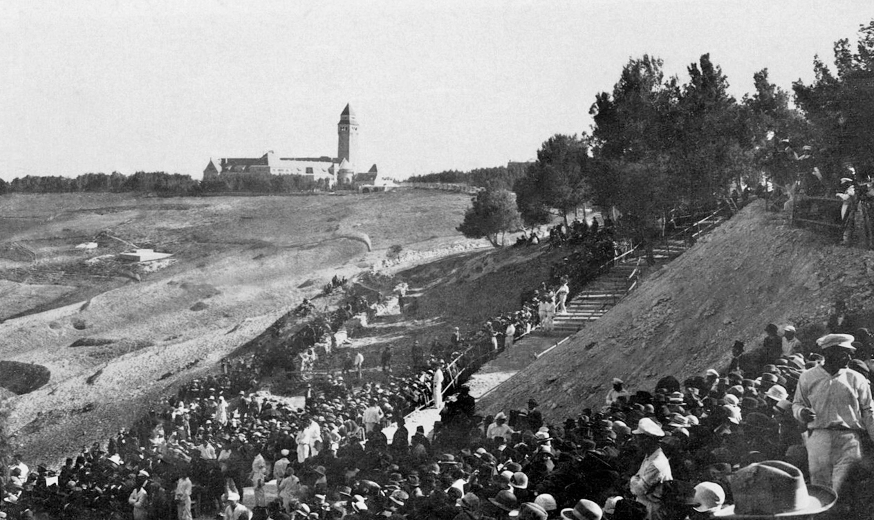 Black and white image of a masonry building in the distance with a square tower. In the foreground, a crowd is lined up as if for a procession.