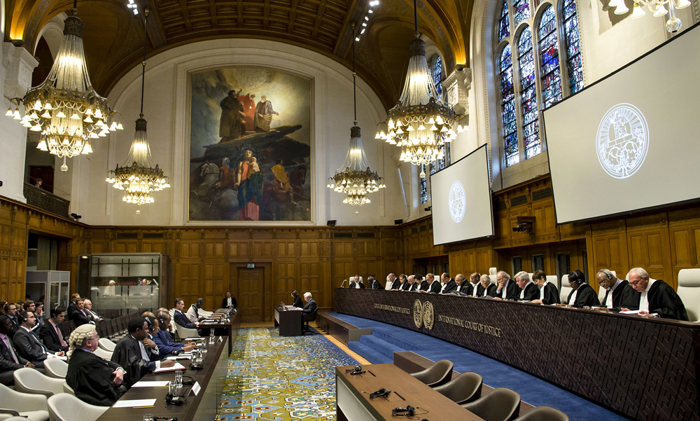 A large wood-paneled room. On the left is a small gallery of spectators and a lawyer in a white wig. Facing them on the right is a bank of almost twenty judges behind a long desk.