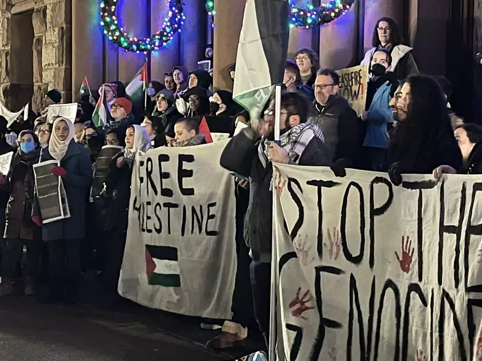 Four rows of protesters under lighted Christmas wreaths hold Palestinian flags and banners reading Free Palestine and Stop the Genocide.