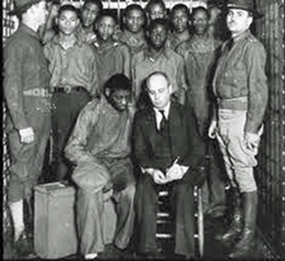 A black and white photo of prison guards flanking a number of Black men. One Black man sits in the foreground next to a white, older man.