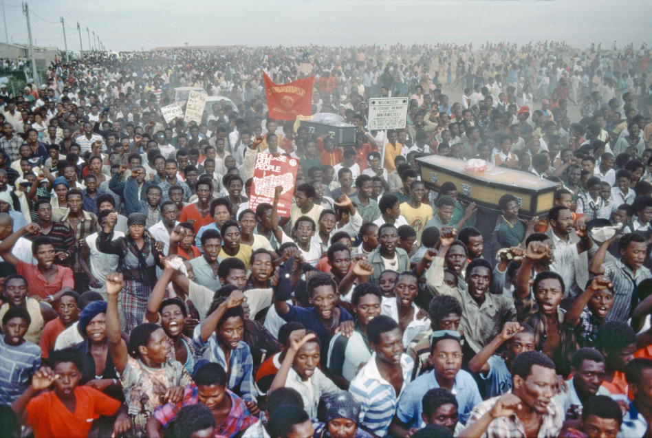 A tightly packed crowd of Black people—as far as the eye can see—escorts two coffins. Many have their fists raised and their mouths open as if chanting or singing.