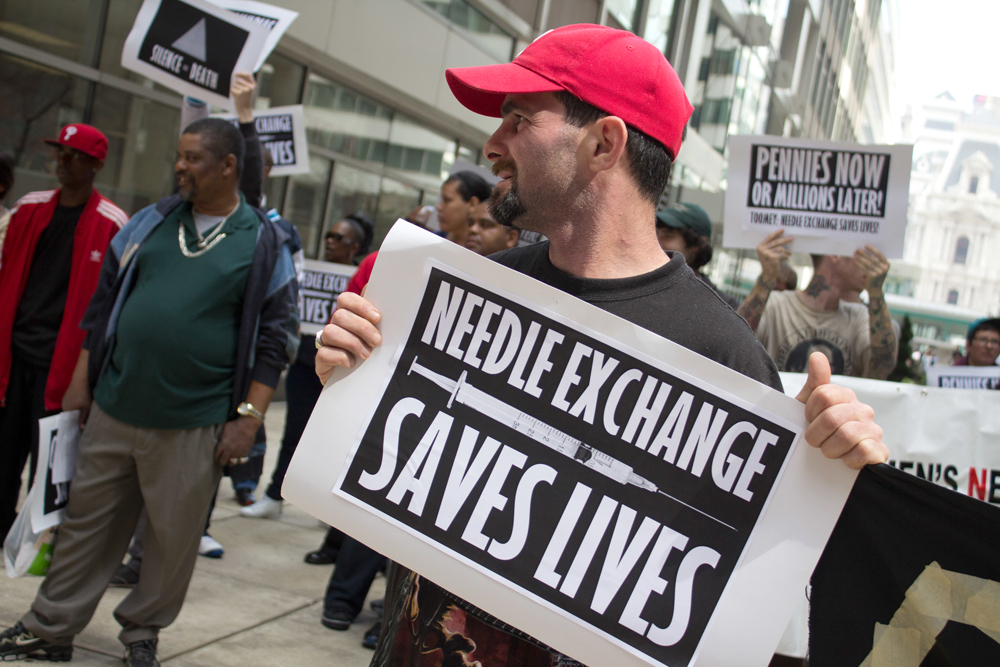 Street demonstrators hold up signs. In the foreground, a man in a red cap holds a sign that says “Needle exchange saves lives.”