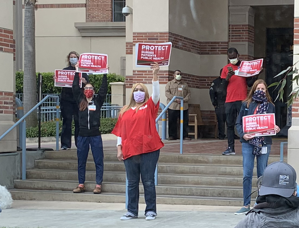 Workers on medical center steps hold signs that say, “Protect nurses, patients, public health.”