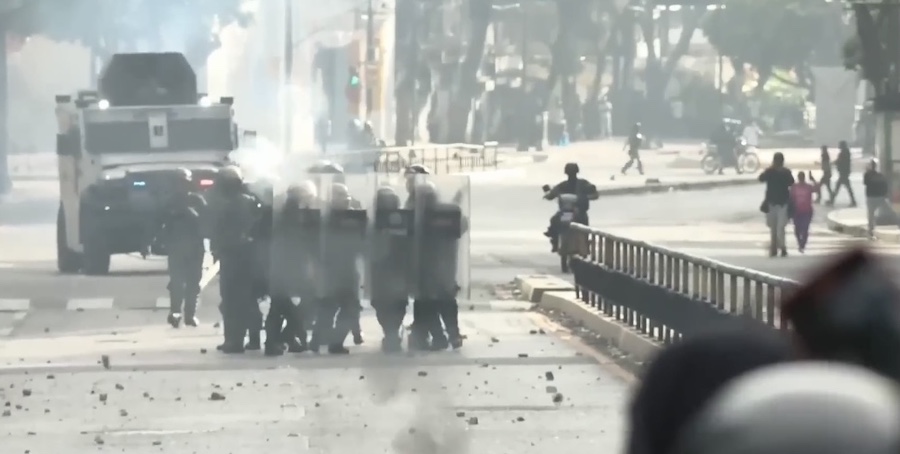 Photo from Venezuela on August 11, 2024. It shows a group of about a dozen riot police behind riot shields and holding automatic weapons, ahead of an armored personnel carrier, all marching towards a group of protestors in the extreme foreground to the right. Photo by Confidencial.