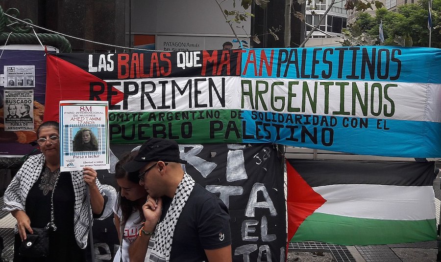 Demonstrators at the annual March in Memory of Truth and Justice in Buenos Aires in 2018. The photo shows a large banner made up of  the Palestine flag on the left and the flag of Argentina on the right. The banner reads: “The bullets that kill Palestinians repress Argentinians” with the name of Committee in Solidarity with the People of Palestine at the bottom in black lettering. A woman in the foreground is wearing a keffiyeh and holding a placard with the image of Ahed Tamimi, a then-17 years old Palestinian political prisoner being held by Israel.  Photo by Gastón Cuello.