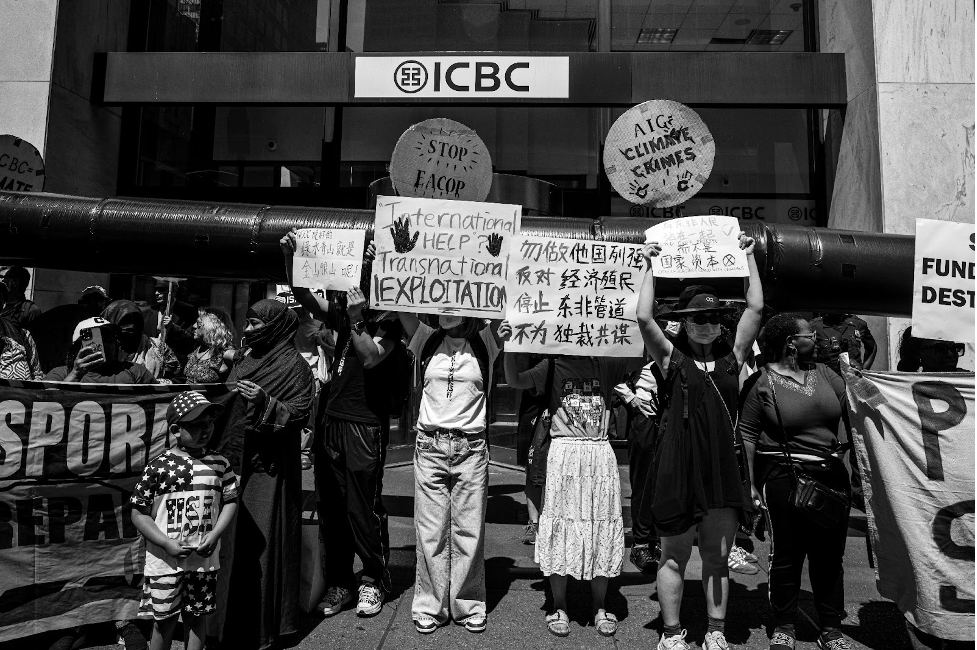 Black and white photo of about a dozen protesters in front of a building whose sign reads ICBC. Protest signs are in English and Chinese; one English sign reads: International Help? Transnational Exploitation.