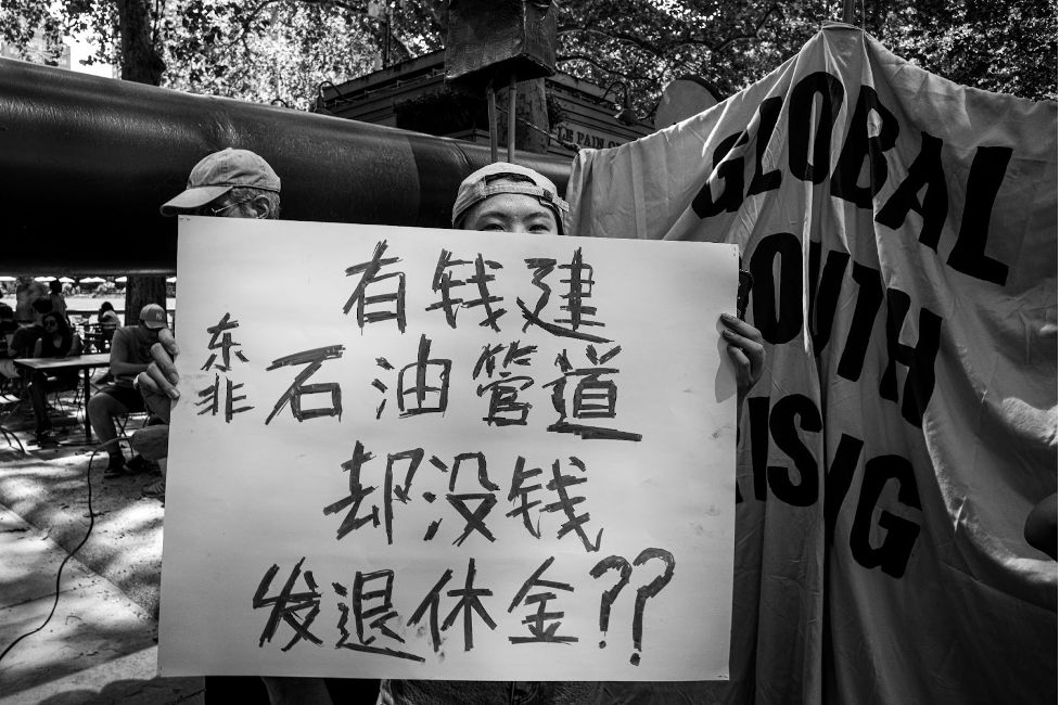 Black and white photo of a protester holding a sign in Chinese. The translation reads “Money for EACOP but no money for retirement?”