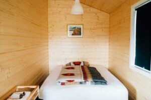 A small, wood paneled bedroom with a bed with a white cover and carefully folded blanket, a picture, and a window.