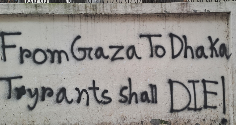 Black graffiti on a dirty white wall says, “From Gaza to Dhaka, tyrants shall die.”