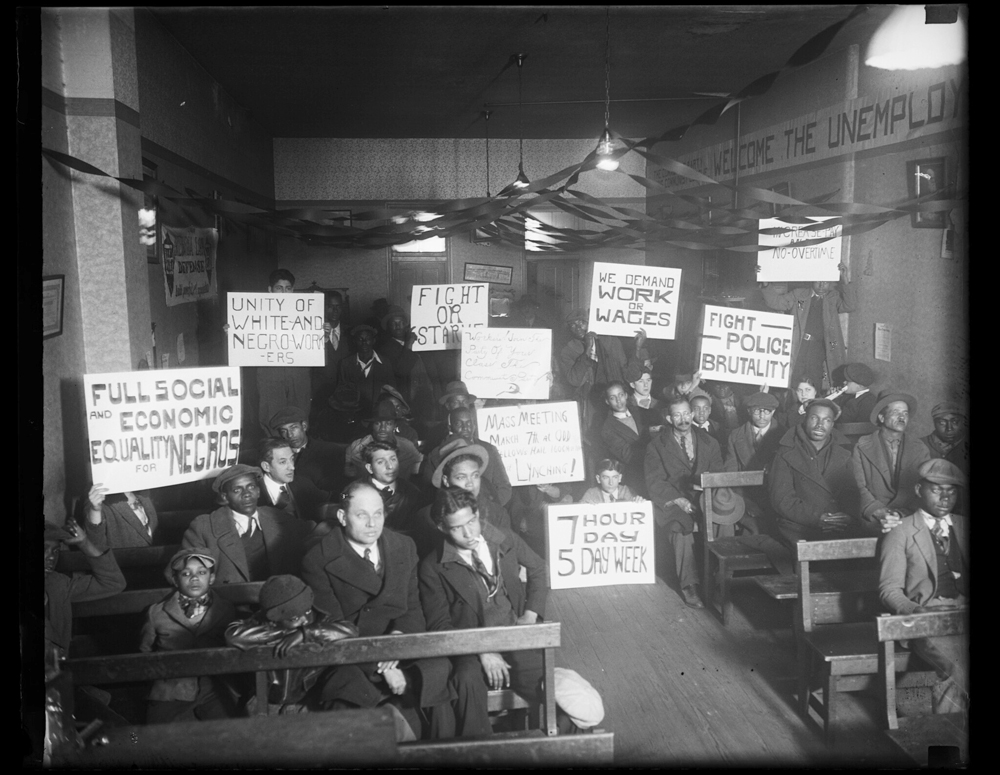 Multiracial crowd of men at a dimly-lit indoor rally holding signs. Black and white image.