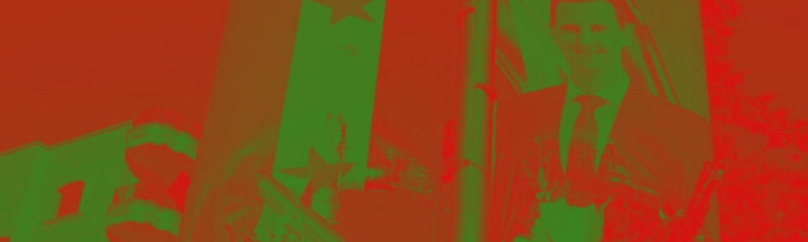 A photo of a pole with the Syrian flag hanging next to a picture of Bashar al-Assad. Behind it is a building. Heavy green and red filter over the whole photo.