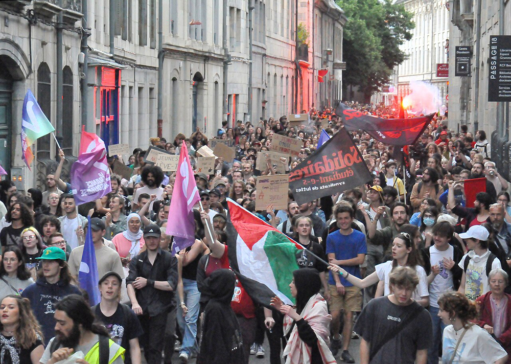 Young crowd approaching the viewer down a fairly narrow city street carrying banners and flags, including a Palestinian flag.