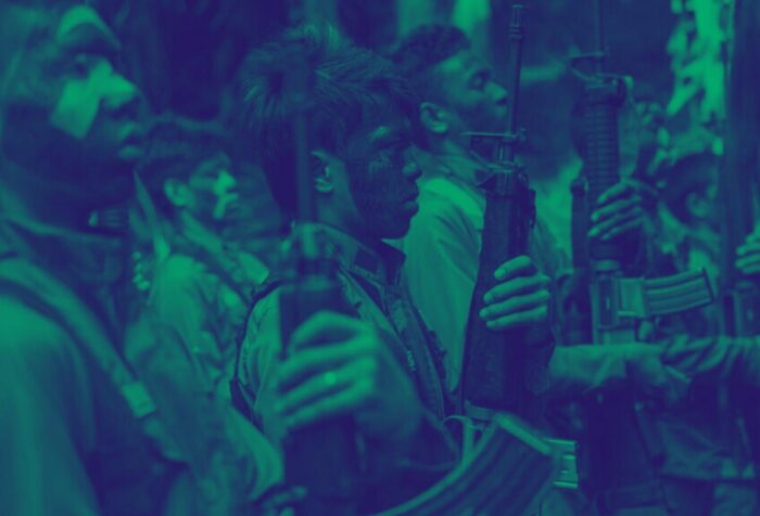 Photo of young men holding guns with their faces painted stand in lines and face right. A dark blue filter over the photo.