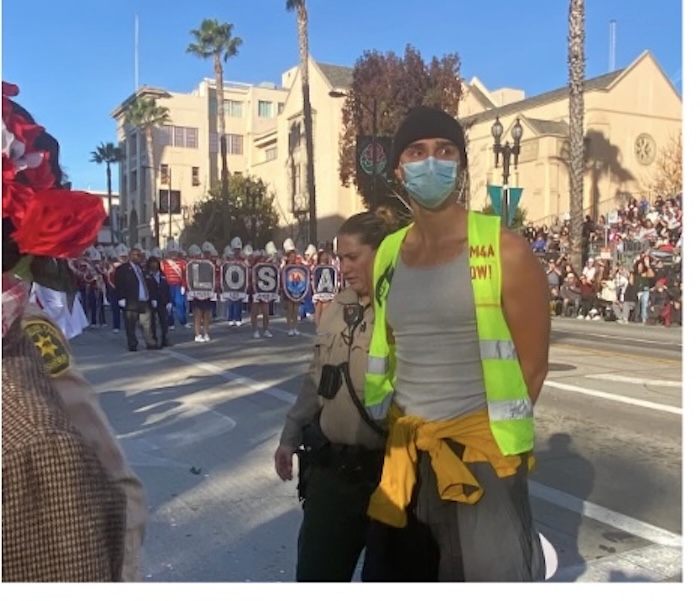 Palestine solidarity march participant, under arrest at the Rose Parade. Participant wearing a fluorescent yellow safety vest and mask with hands handcuffed behind his back being led from behind by a police office in tan and brown. In the background are the march participants carrying the Palestine banner (not legible). Photo by Teto. 