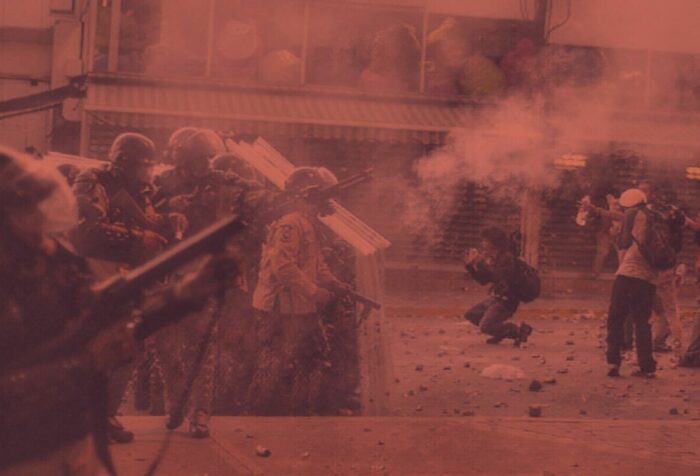 Militarized police in headgear aim guns toward a group of students in the street, surrounded by wafts of teargas.